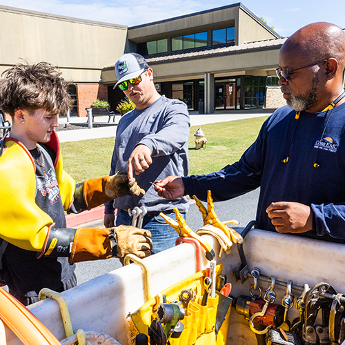 Two linemen standing around a bucket truck bucket with a student wearing rubber gloves